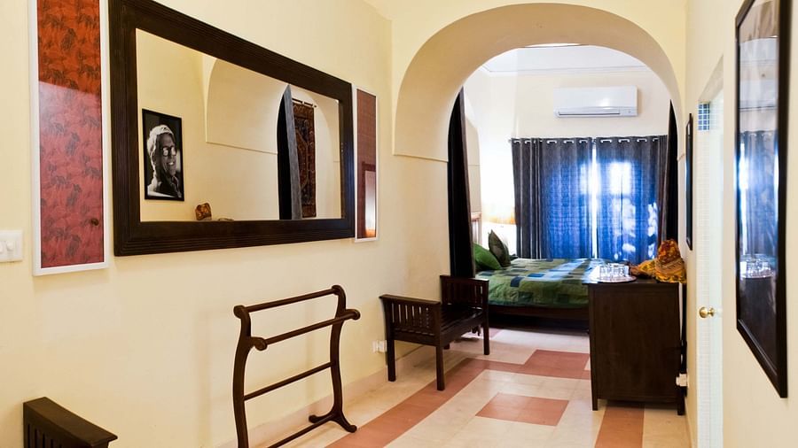 Hotel hallway with a large mirror, artwork on the walls, a towel rack, and a view into a bedroom with blue curtains at Tijara Fort-Palace, Alwar.