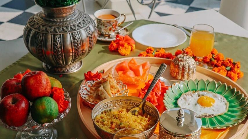 A breakfast spread laid out on a table on a balcony, featuring fruits, eggs, drinks, and other dishes, with a view of blue railings and greenery | Dileep Kothi, Jaipur
