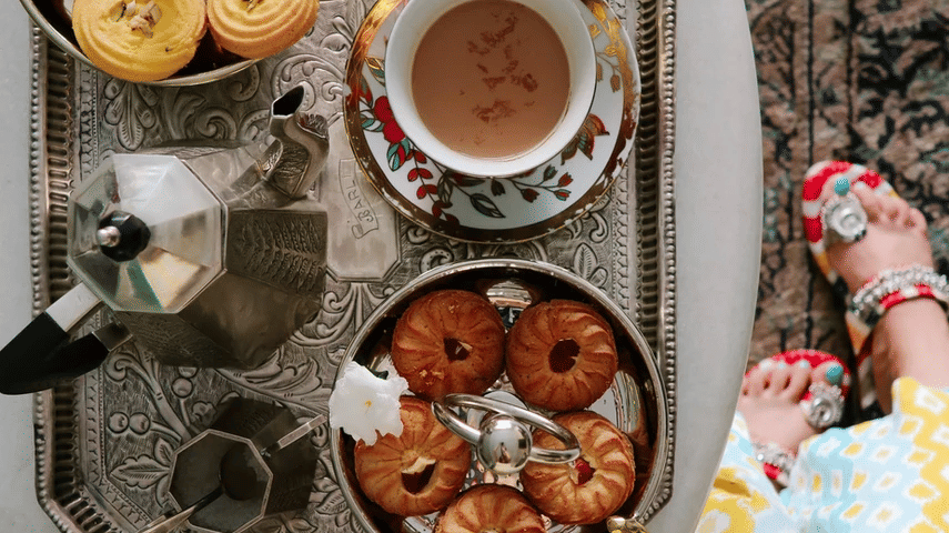 A flat lay of a silver tray with coffee, pastries, and a yellow flower on a white table, with a person's patterned dress and feet visible at the edge | Dileep Kothi, Jaipur