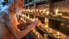 An elderly priest is lighting numerous small oil lamps arranged in rows within a temple or religious building.