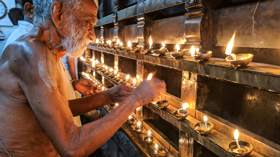 An elderly priest is lighting numerous small oil lamps arranged in rows within a temple or religious building.