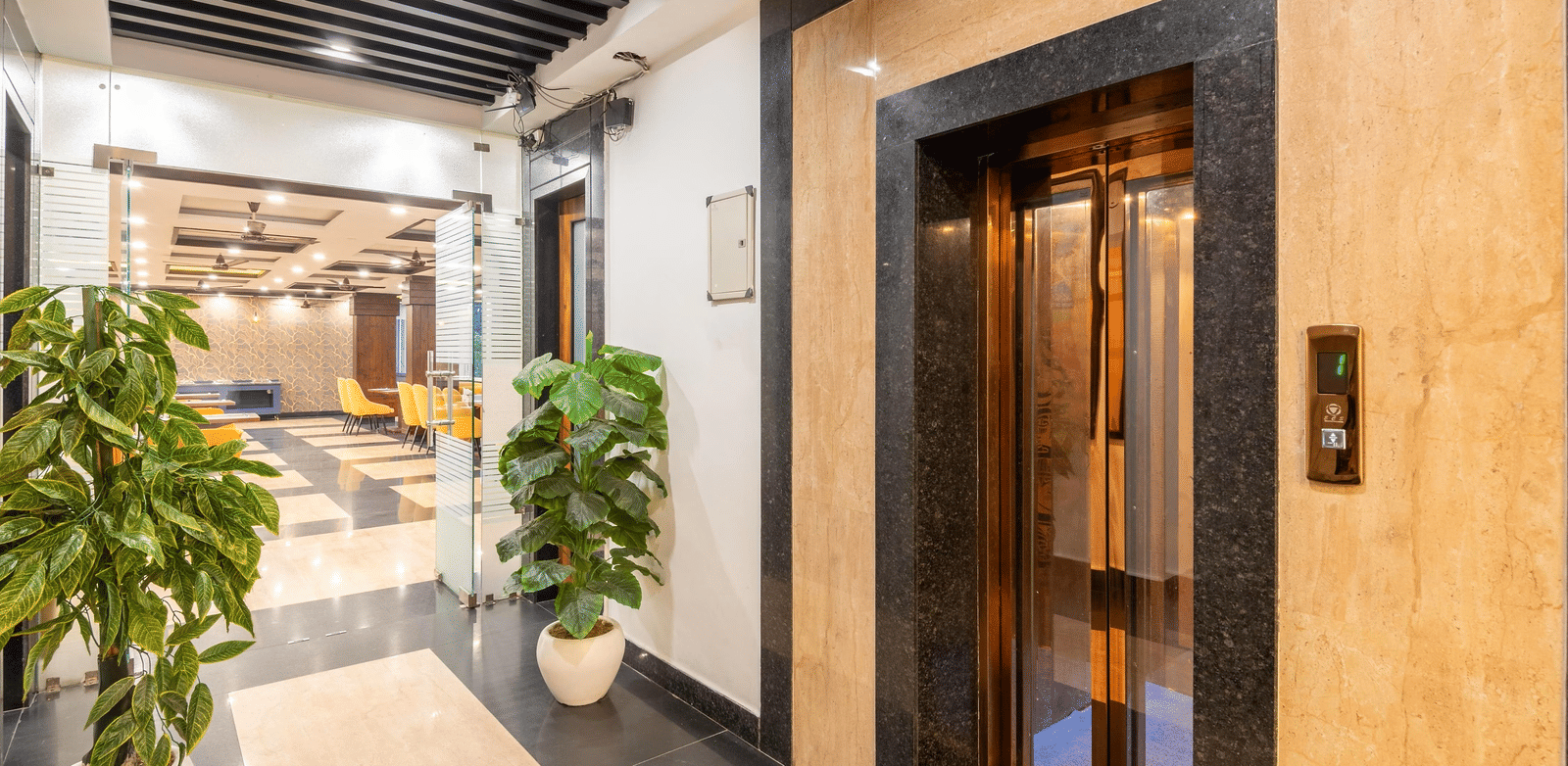 A hotel lobby hallway featuring a glass elevator and potted plants at Perfectstayz Premium Shanti Heritage, Haridwar