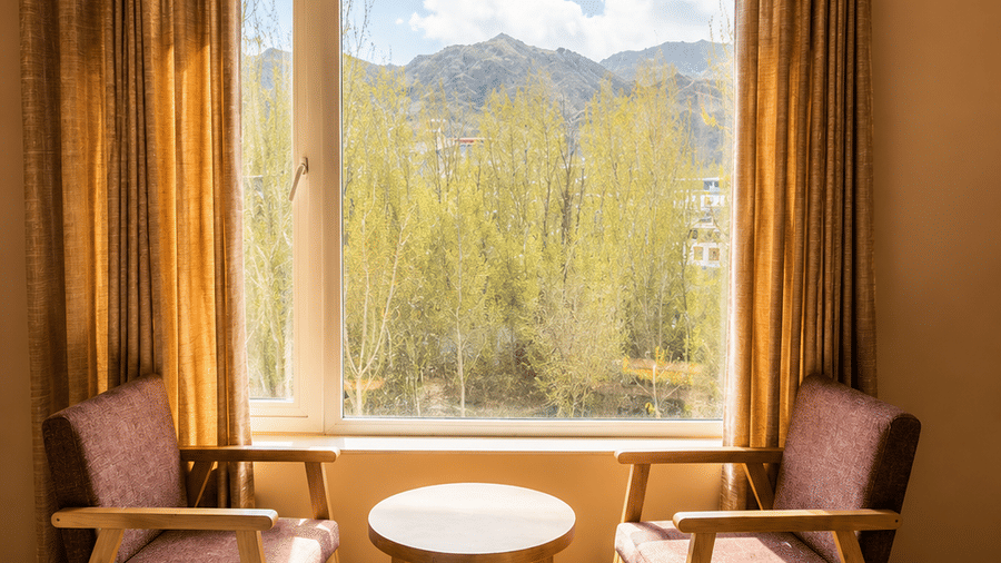 Hotel room seating corner with two wooden armchairs, a round table and a large window overlooking snow-capped mountains and greenery outside.