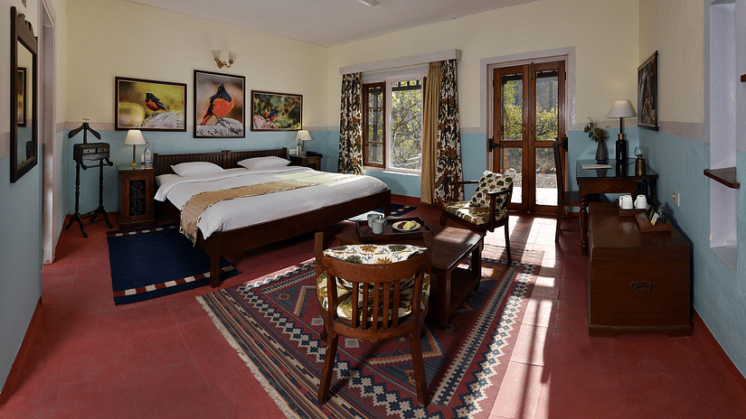 A spacious and elegantly decorated room featuring a large bed, vintage wooden furniture, and vibrant artwork on the walls. Sunlight streams through the windows, highlighting the cosy seating area and colourful rugs.