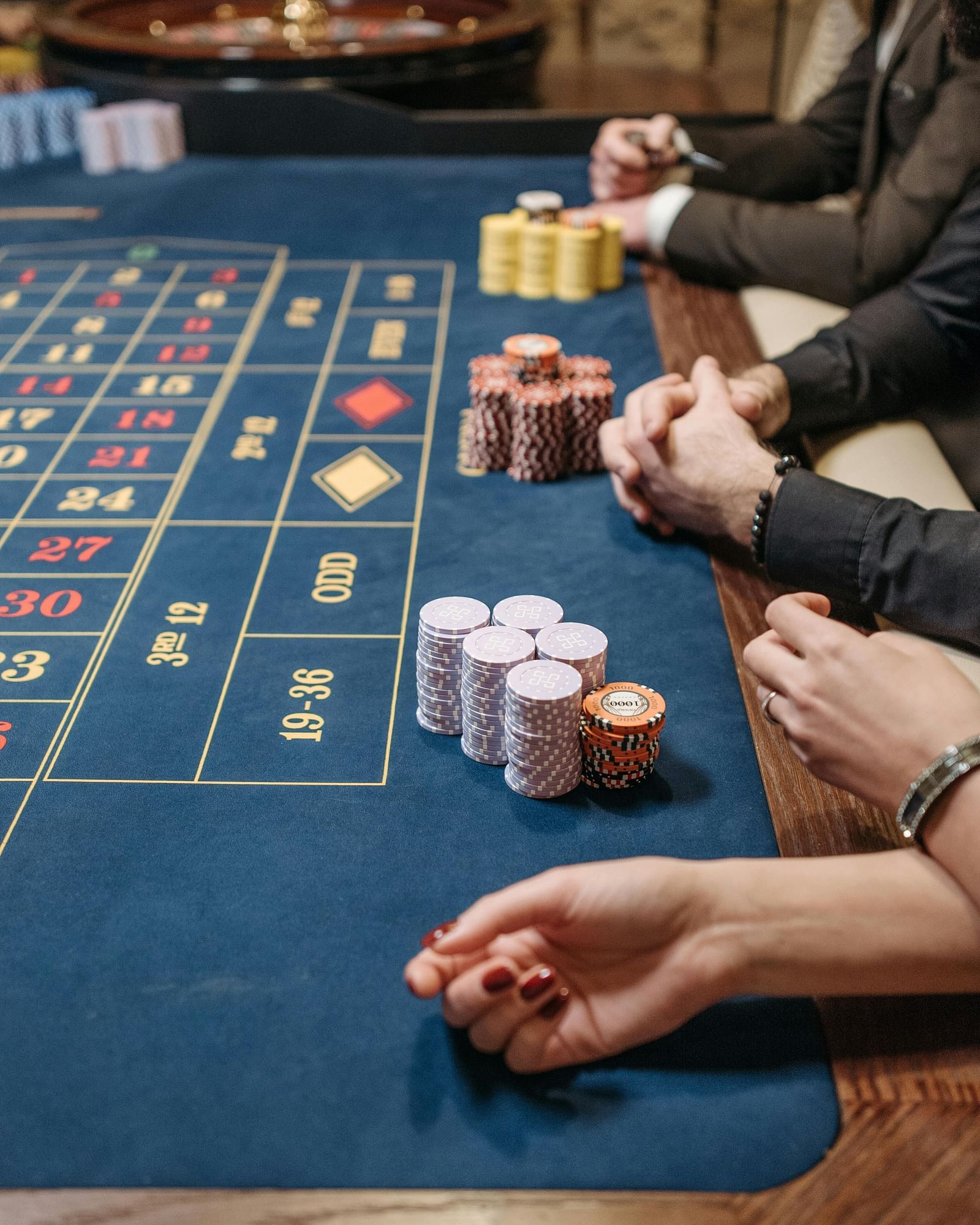A blurred close-up shows people at a roulette table, hands resting near stacks of chips on the blue felt surface.