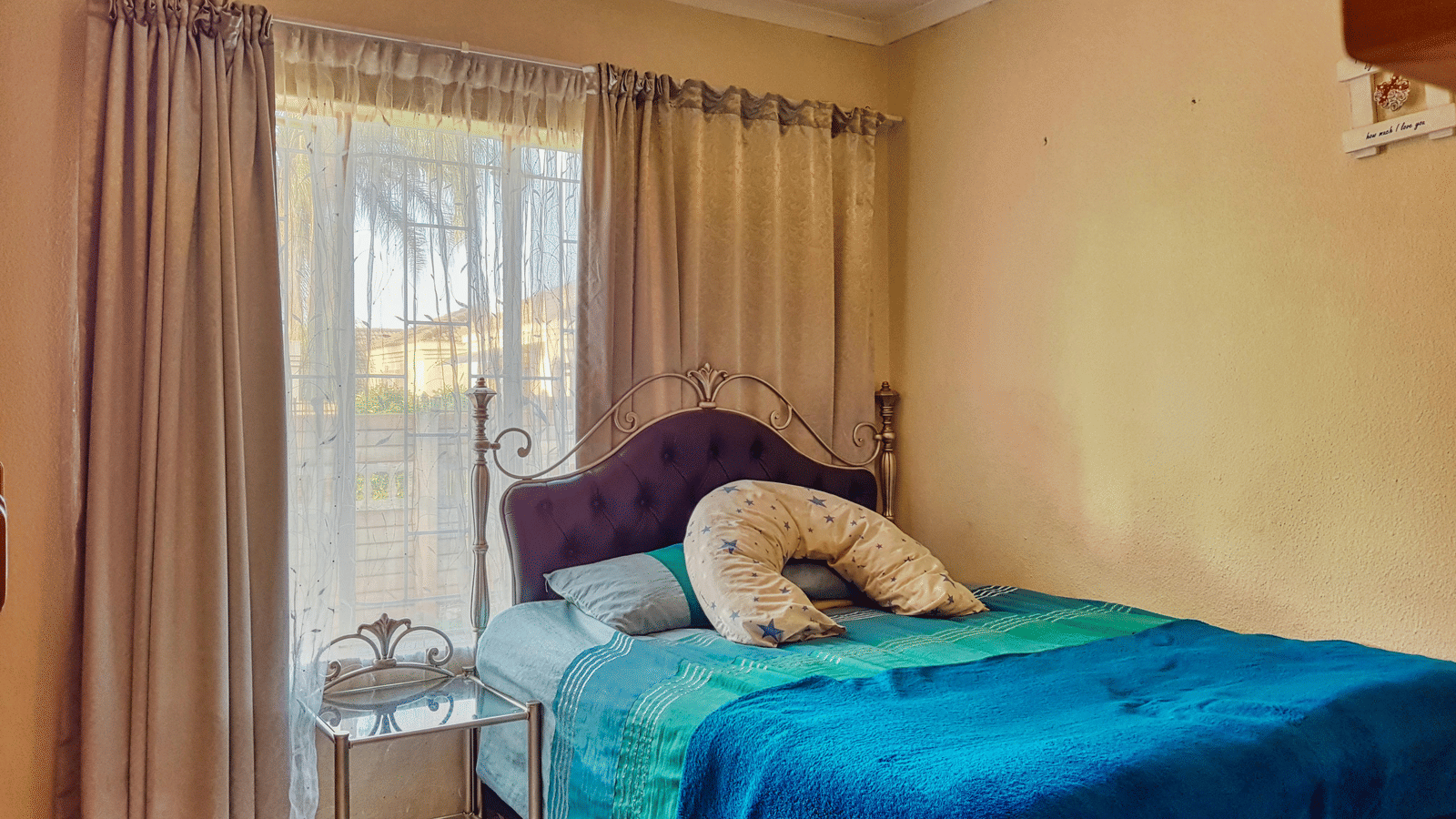 A bedroom featuring a double bed with blue sheets spread neatly over it with beige curtains covering the window behind it.
