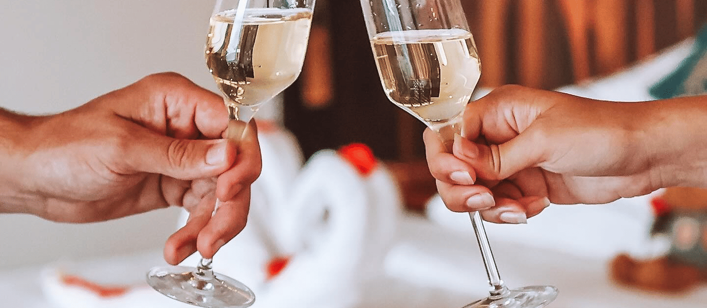Two people bringing their champagne glasses together in a bedroom where the bed is decorated with flower petals and towels.