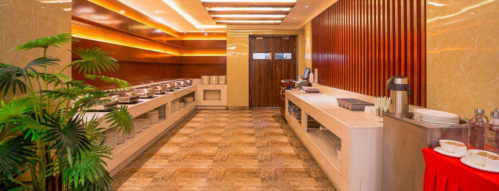 A bright, long hallway featuring a buffet counter with a red tablecloth and green potted plants, leading towards a dining area at Mansha Hotel Regency.