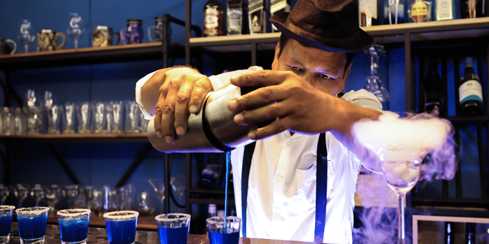 A bartender in a hat and braces adding dry ice to blue cocktails, creating a smoky, theatrical effect at Hotel Hukam's Lalit Mahal.