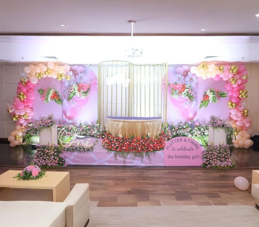 A decorated hall in Dhanalakshmi Srinivasan Hotels, Perambalur, set up for a celebration with a stage adorned with pink and gold balloons, flowers, and a banner. There are sofas and a coffee table in the foreground.