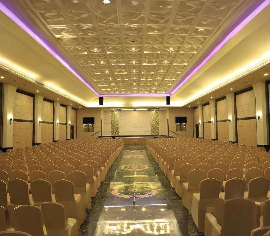 A large empty hall in Dhanalakshmi Srinivasan Hotels, Perambalur, with rows of chairs facing a stage area. The hall has dark marble flooring and a patterned ceiling with lighting.