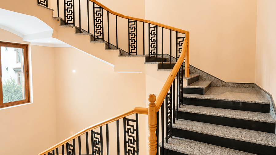Staircase with a wooden handrail and metal balustrade, spiralling upward with patterned flooring and natural light from a nearby window nearby.