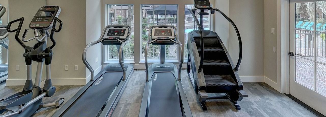 Indoor gym room with four cardio machines featuring two treadmills, a cross-trainer, and a stationary bike, facing tall arched windows.