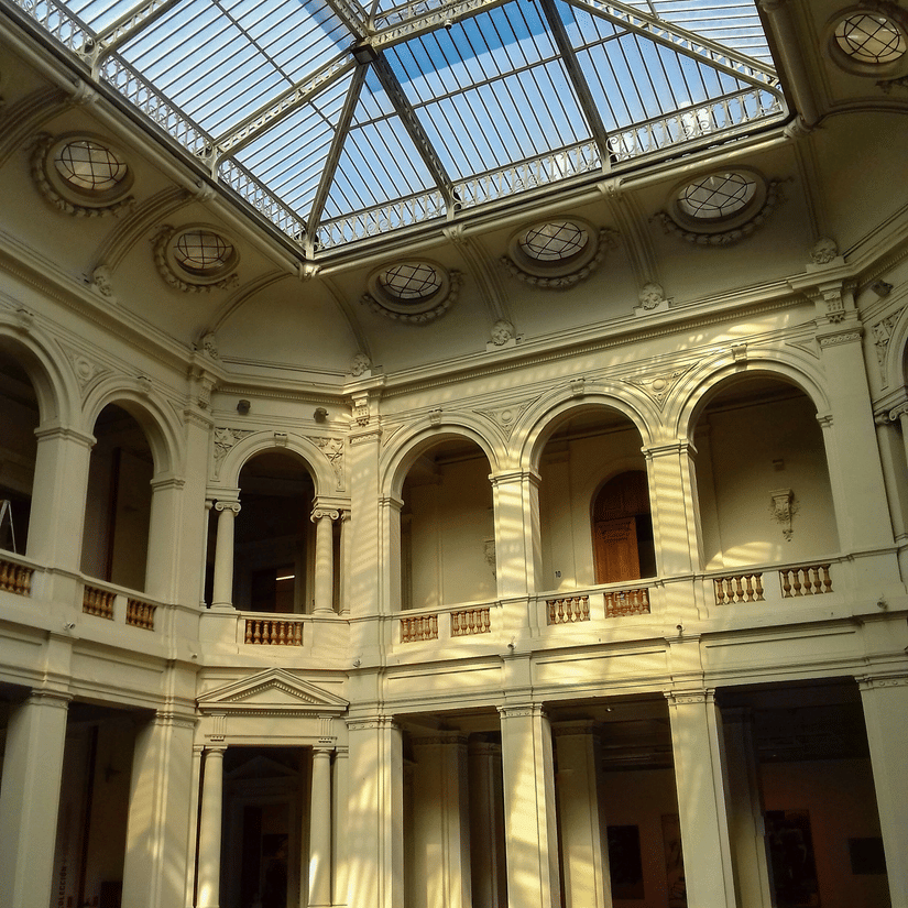 Sunlight streams through a grand glass atrium ceiling into a classical courtyard with arched stone balconies.