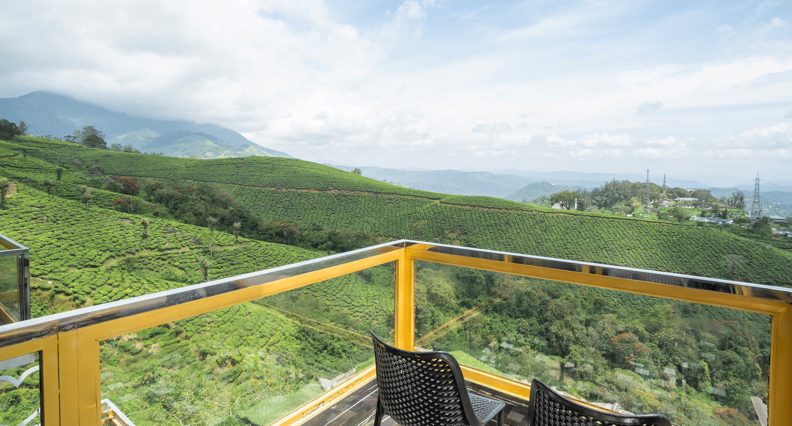 The private balcony of the Jacuzzi 180-degree Suite at Parakkat Nature Hotel and Resorts, Munnar, featuring a glass railing, a small table, and an expansive view of the rolling, terraced tea estates under a sunny sky.