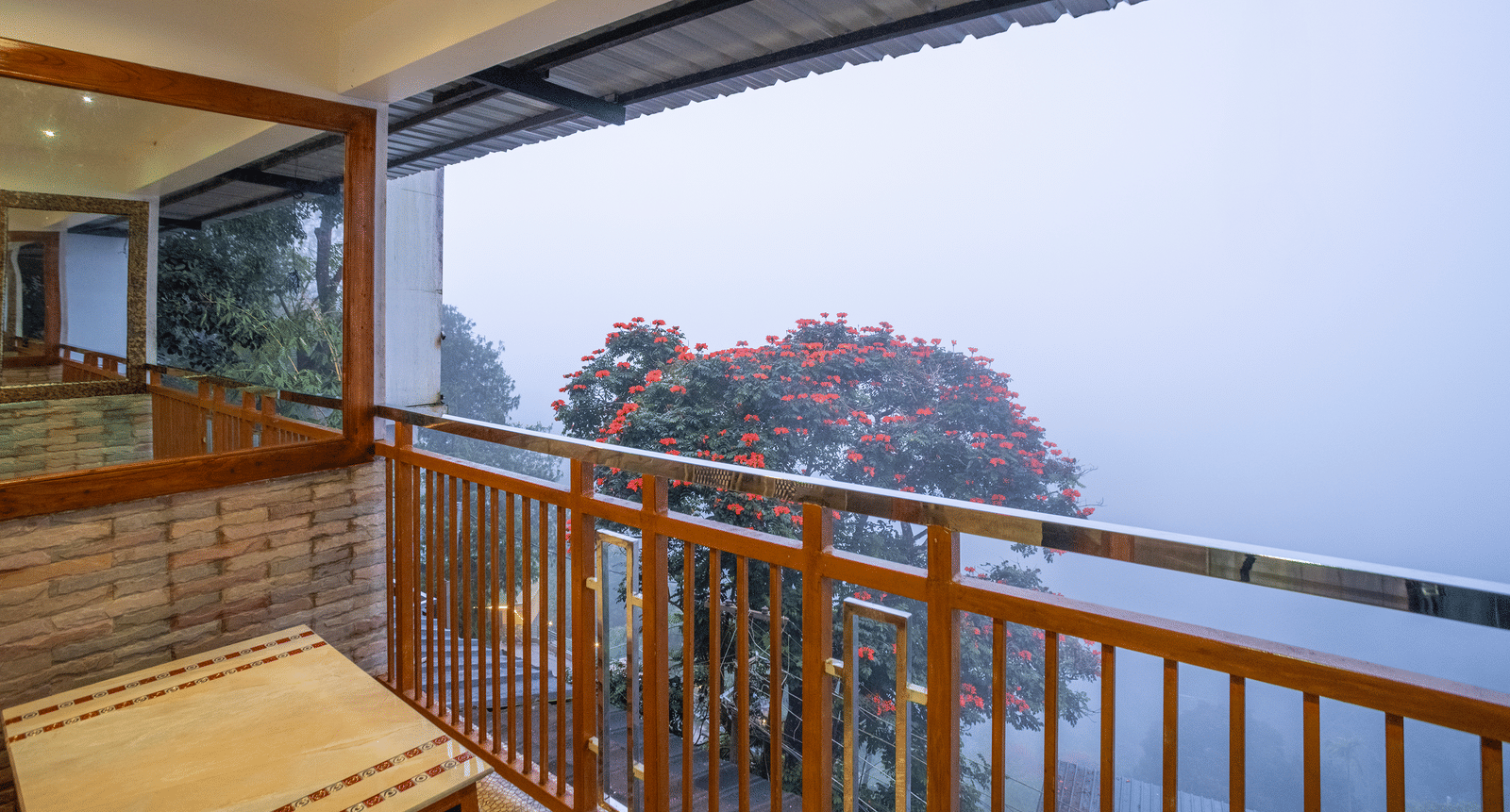 The balcony of the Premium Club Suite at Parakkat Nature Hotel and Resorts, Munnar, featuring a wooden railing, a glass partition, and a scenic, foggy view of the surrounding hills.
