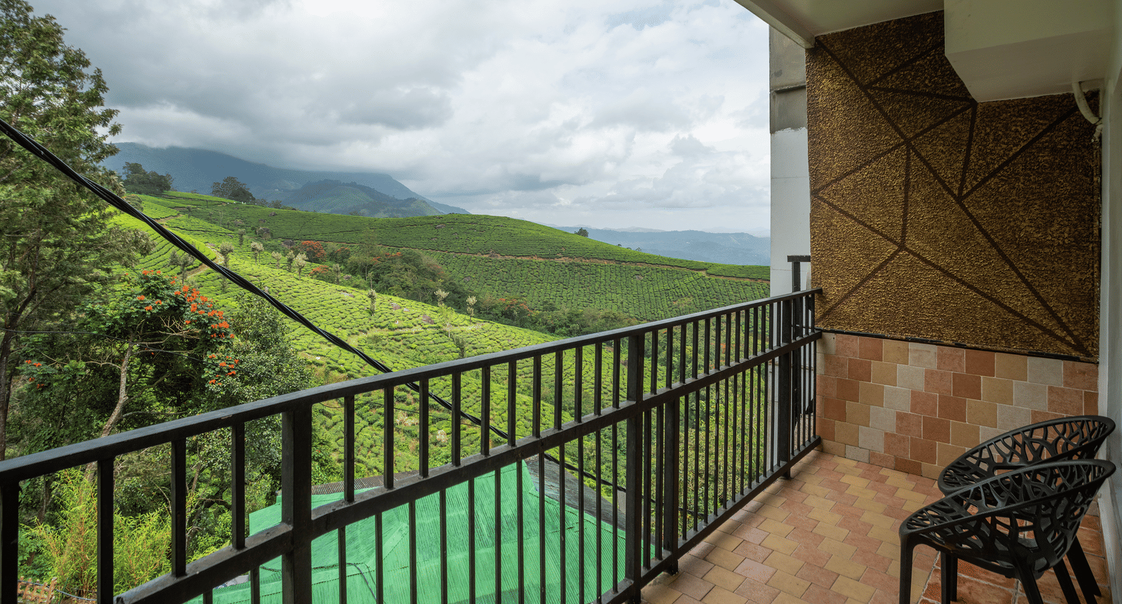 The private balcony of Premium Room at Parakkat Nature Hotel and Resorts, Munnar featuring a metal railing and seating area overlooking greenery under a cloudy sky.