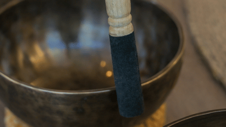A hand holding a wooden mallet to play a metal Tibetan singing bowl for sound healing therapy at Kavya Himalayas.