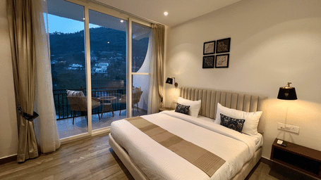 A bedroom featuring a bed with pillows, wall lamps, and glass doors leading to a balcony with hill views at Ridge By The Lake Hill, Bhimtal.