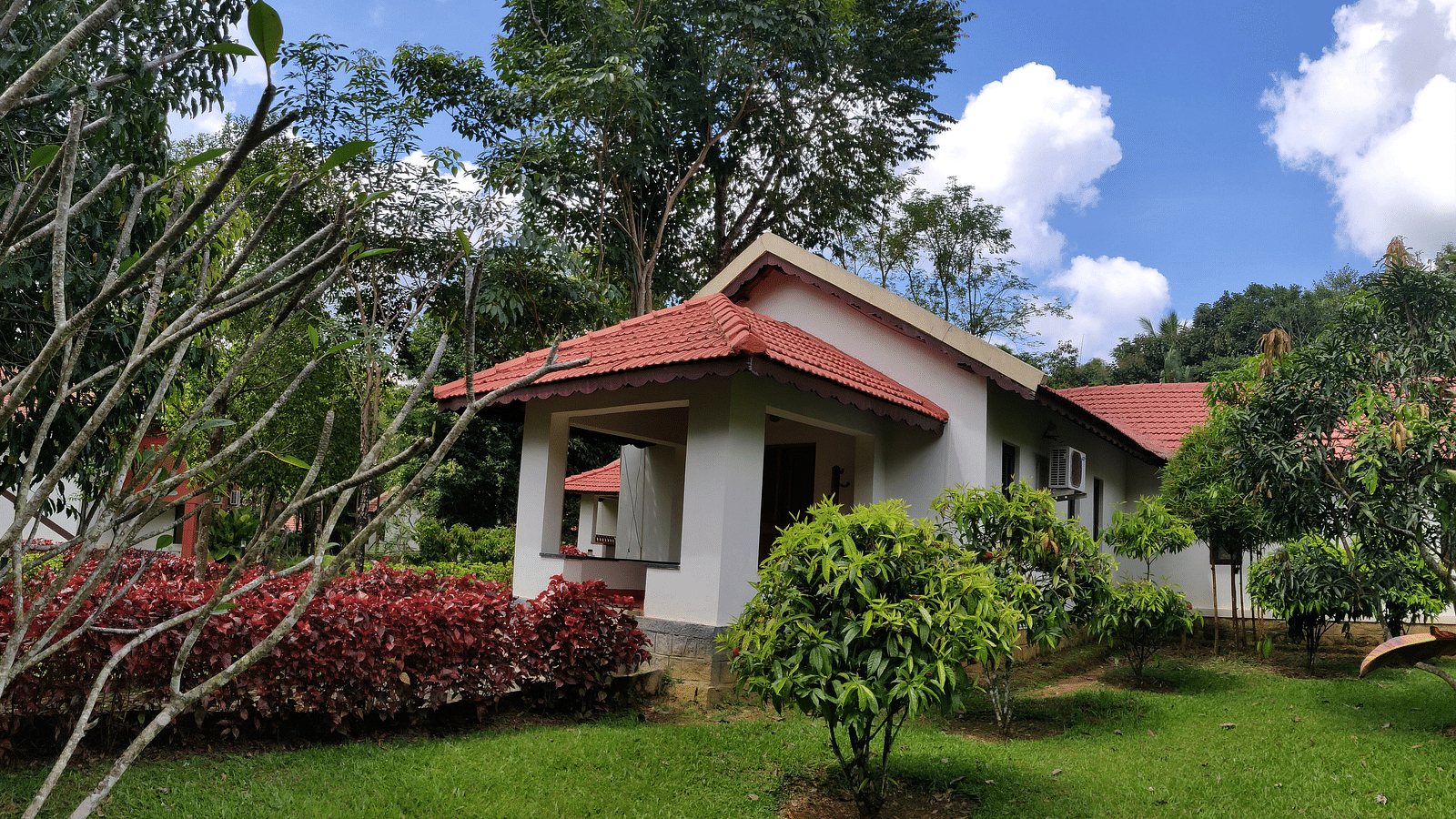 A single-story cottage with a red tiled roof and white walls surrounded by a lush garden with red-flowered shrubs, green lawns, and tall trees against a blue sky with fluffy clouds.
