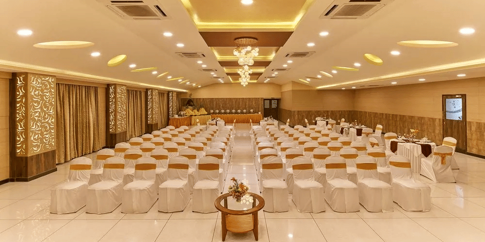 A banquet hall is arranged in theatre-style seating with rows of white-clothed chairs featuring gold sashes, brightly illuminated by a modern chandelier and recessed ceiling lights at Ramanashree Richmond.