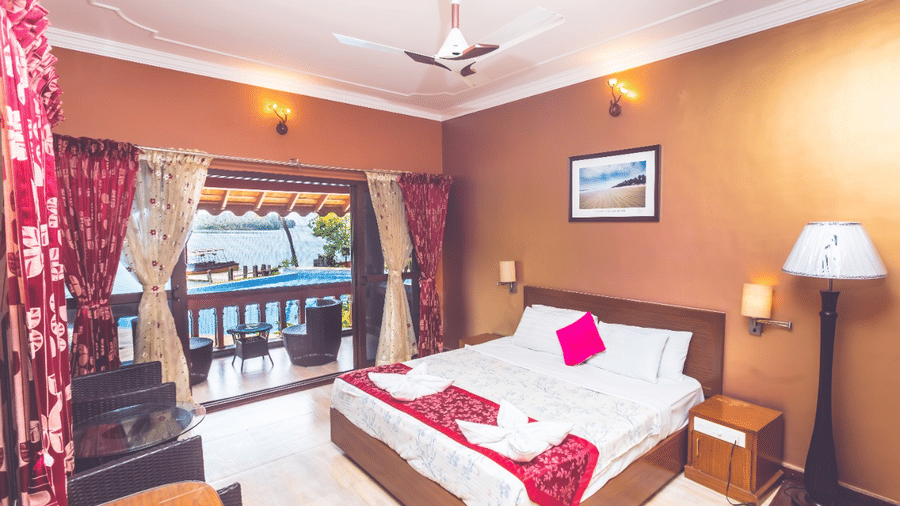 A brightly lit room with a queen bed and pink floral accents, wall art and a window overlooking the water at Paradise Lagoon Resort, Udupi.