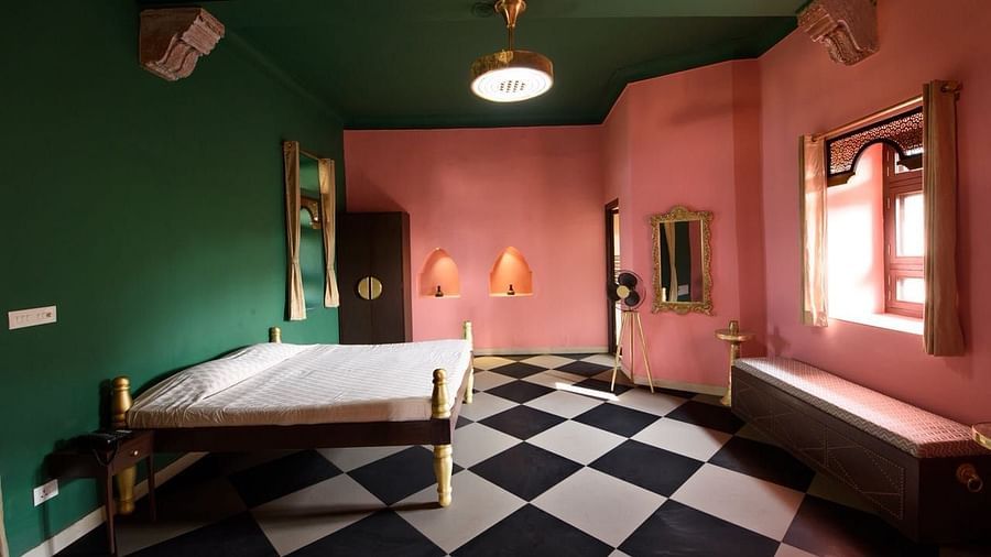 A guest room at Tijara Fort-Palace - 19th Century, Alwar features a bed, a mirror, a standing fan, a door with curtains, and checkered flooring.