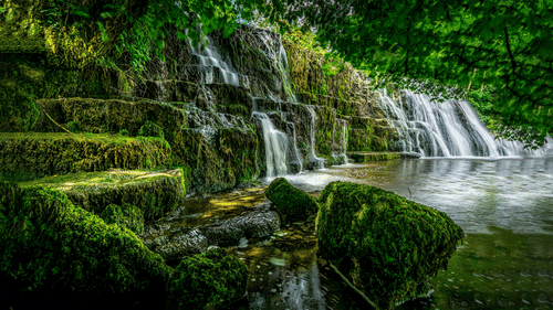 An overview of a Waterfall cascading down to a water body with greenery surrounding it