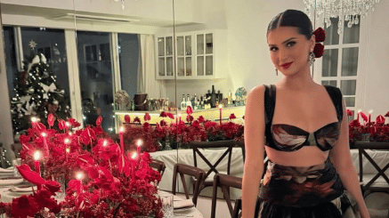 Actress Tara Sutaria posing in a patterned black two-piece outfit next to a festive dining table filled with red floral arrangements and candles, with a Christmas tree in the background.