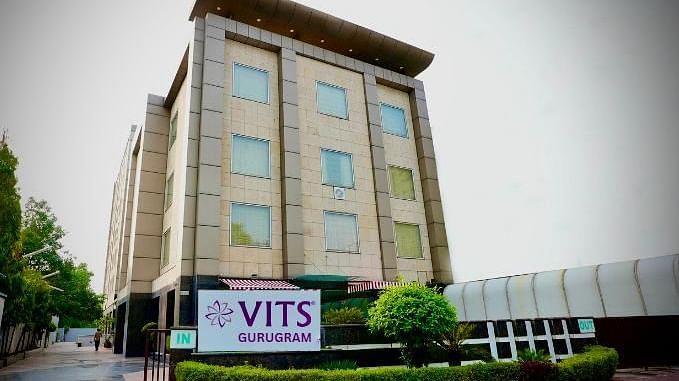 A low-angle exterior shot of a multi-storey building with a sign that reads VITS GURUGRAM, surrounded by greenery