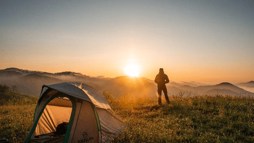 a man standing next to a tent relishing the sunrise under a clear sky 