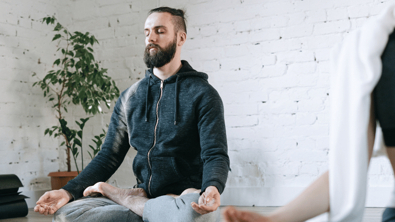A person sitting cross-legged on a yoga mat with eyes closed, meditating in a calm indoor space with a plant and white brick wall in the background.