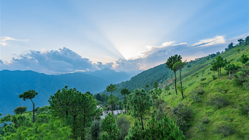 A view of a lush green valley with the sun shining through the clouds in the distant horizon.