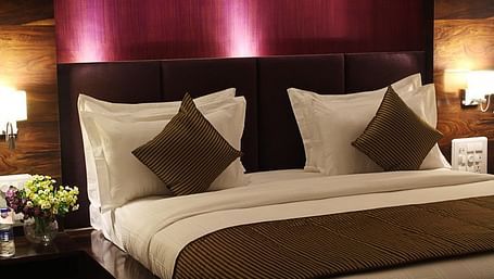 A warmly lit hotel room showcases a king size bed with a prominent dark headboard, crisp white linens, and decorative throw pillows.