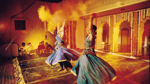 Two dancers in swirling blue and green skirts perform with lit torches at Piramal Haveli, while musicians and onlookers are seated in the background.