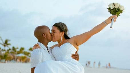 A couple celebrating on a beach, with the groom carrying the bride in his arms against a backdrop of the ocean.
