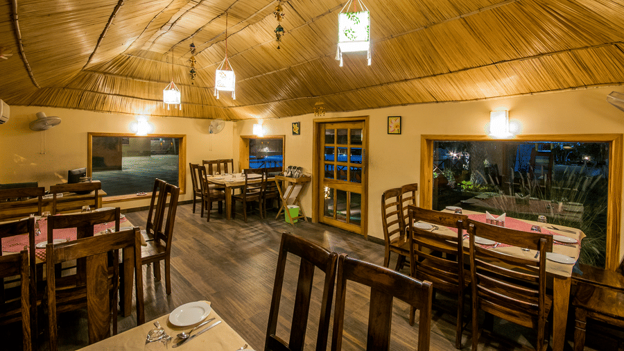 Multiple wooden tables and chairs are arranged inside a dining room with a slatted bamboo-like ceiling at Garh Govind.