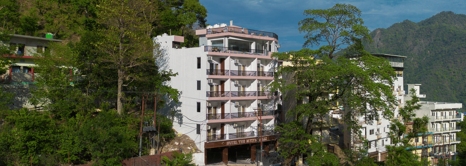 An exterior of a building with multiple storeys, balconies, and windows, surrounded by trees on a hillside in Perfectstayz Premium Mystic Falls, Rishikesh.