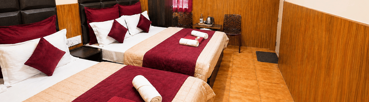 Deluxe Quadruple Room at BN Tourist Home, Ooty, featuring 2 plush beds facing a wall-mounted television screen next to a wardrobe.