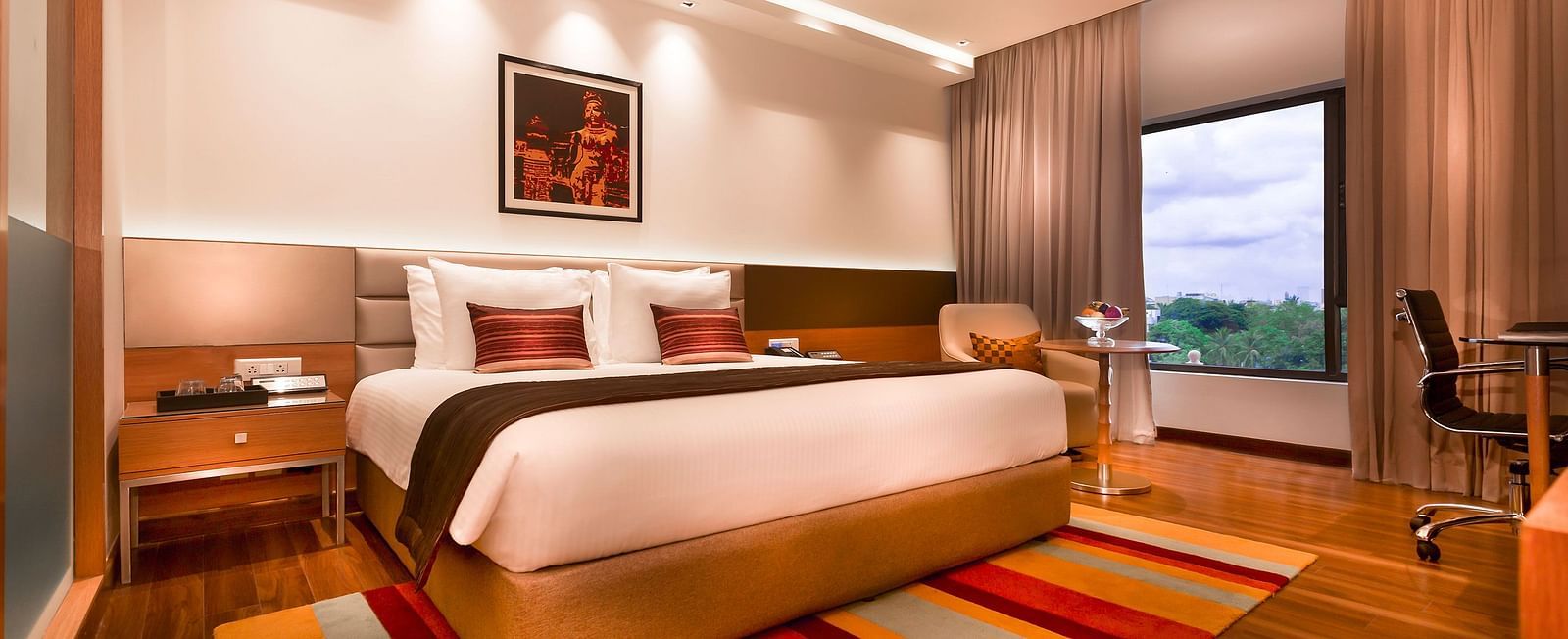 A modern hotel room featuring a king size bed, warm lighting, wooden flooring, a striped rug, and a large window with a view - Grand Chennai by GRT Hotels