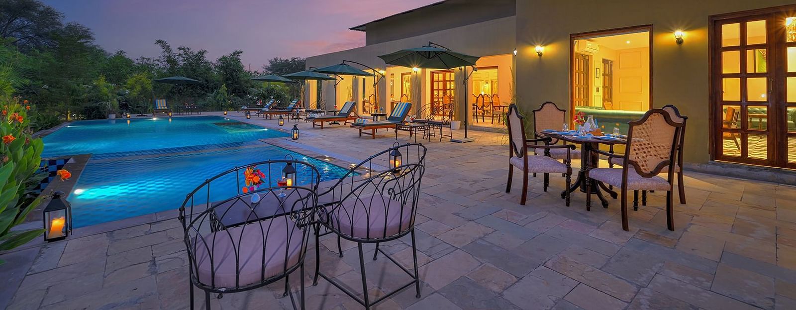 Poolside seating area with cushioned chairs and dining tables, surrounded by greenery and sun loungers under large umbrellas, illuminated by lanterns at dusk - Singhvilas, Ranthambore