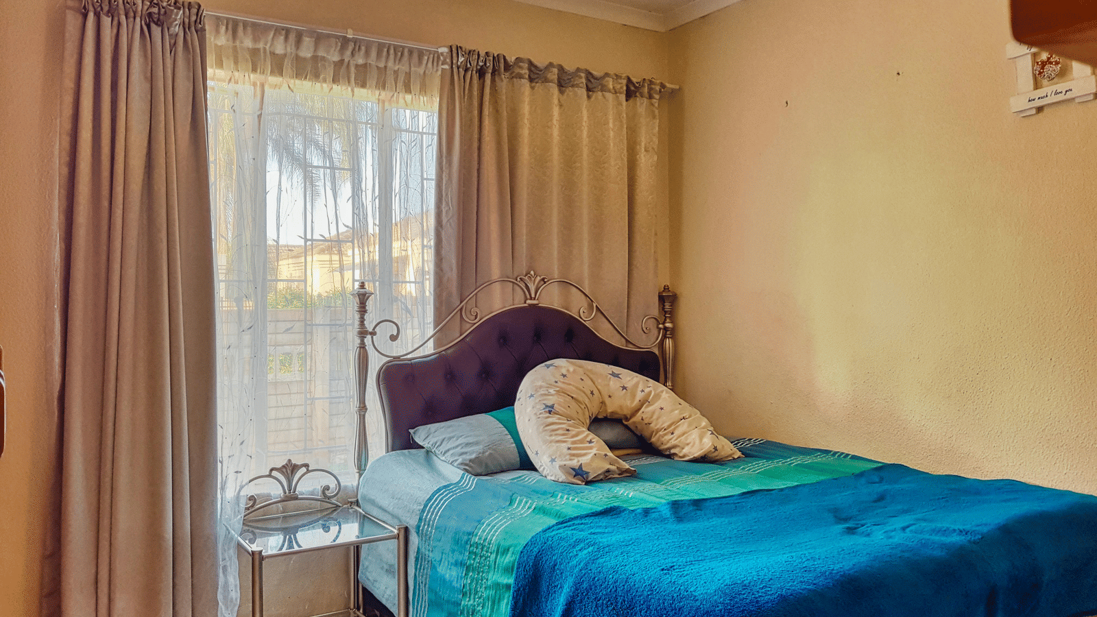 A bedroom featuring a double bed with blue sheets spread neatly over it with beige curtains covering the window behind it.
