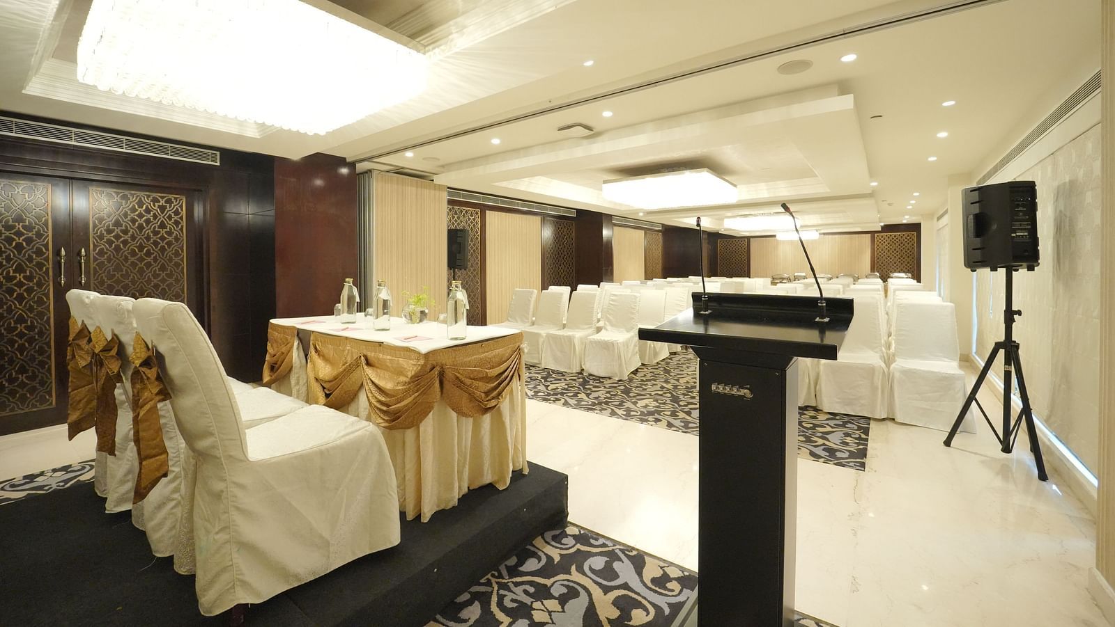 A conference room setup with a podium, speaker system, and chairs with white covers and gold sashes - Fragrant Nature Cochin