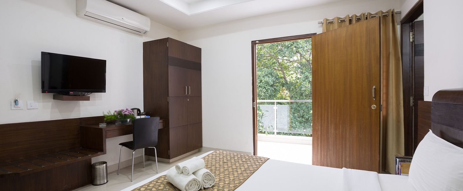 A room with a double bed, a work desk and a chair, wardrobe, and an open door to the balcony - Sanctum Suites BEL Road, Bangalore