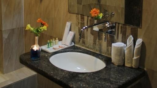 A hotel bathroom with a well-lit mirror, beige tiles and fresh flowers on the vanity counter - TGI Apple Inn, Jaipur 