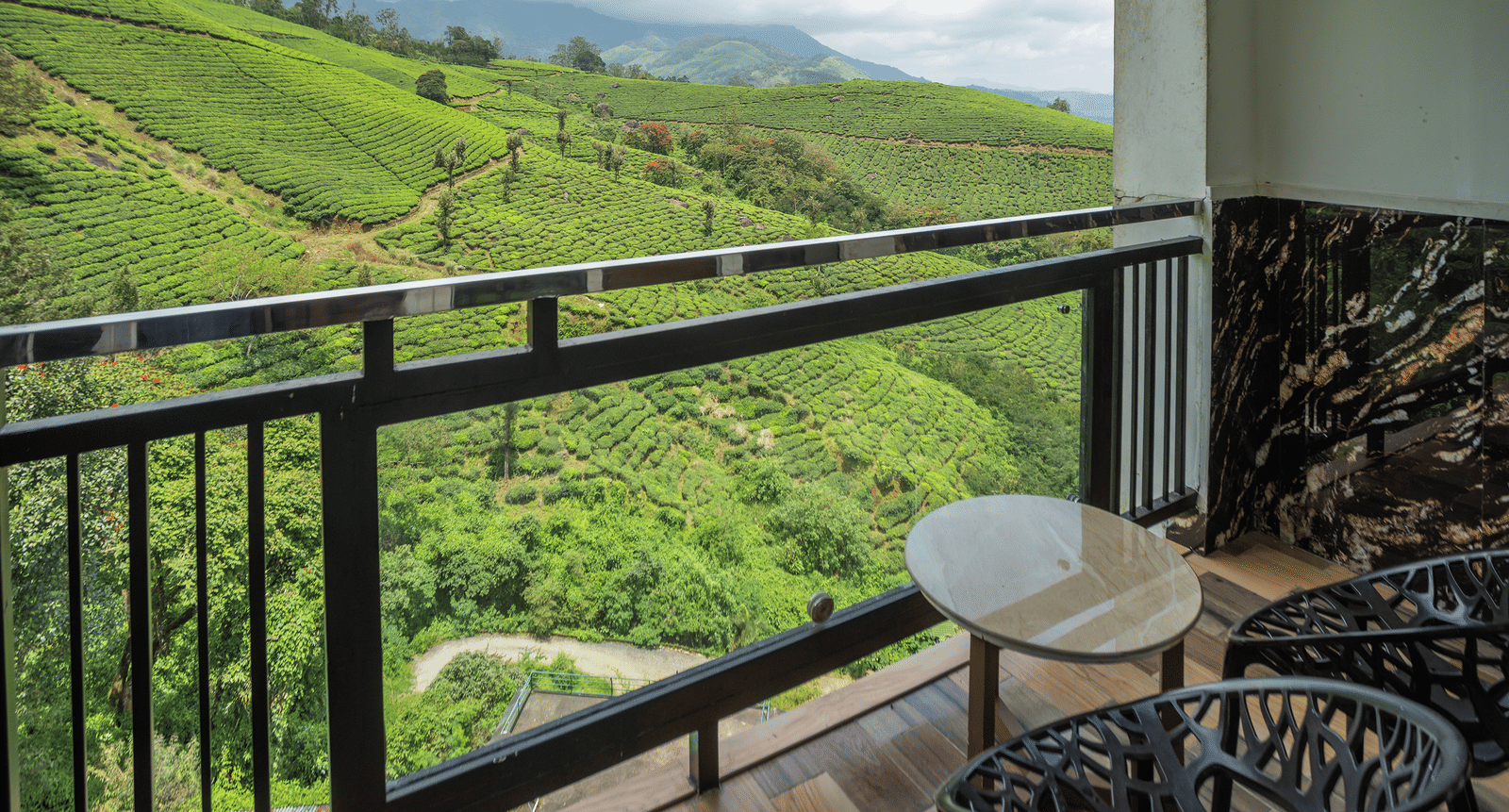 The private balcony of the Honey Moon Suite at Parakkat Nature Hotel and Resorts, Munnar, featuring outdoor seating, a metal railing, and a wide view of the terraced tea estates and distant hills.