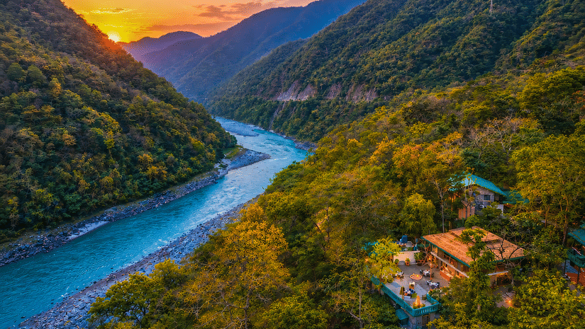 Aerial view of the Neemran's Glasshouse on The Ganges, Rishikesh, surrounded by lush greenery by the river banks, along with a mesmerising sunset in the background.