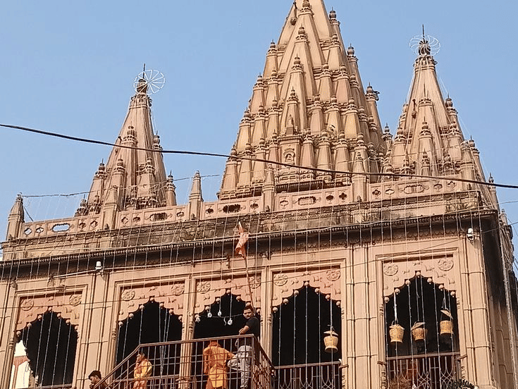 A stone temple with multiple spires located at a river ghat with people standing nearby | HHI Varanasi