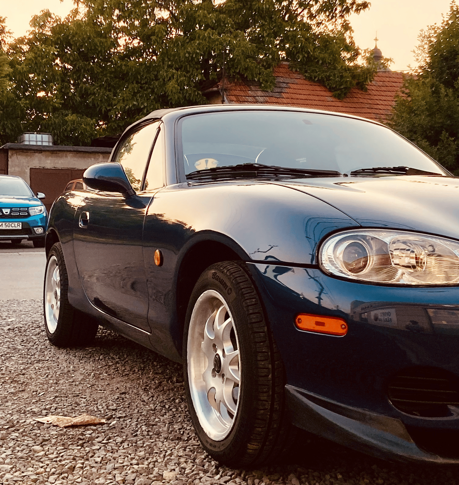 The front end of a low-slung, dark blue convertible sports car parked on gravel, taken during golden hour at sunset.