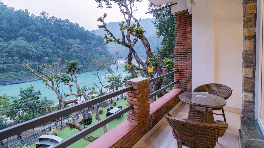 A private balcony featuring a 2-seater round table and 2 chairs, with brick accent walls and metal rails, overlooking a river and trees in the distance | Summit by the Ganges Beach Resort & Spa, Rishikesh